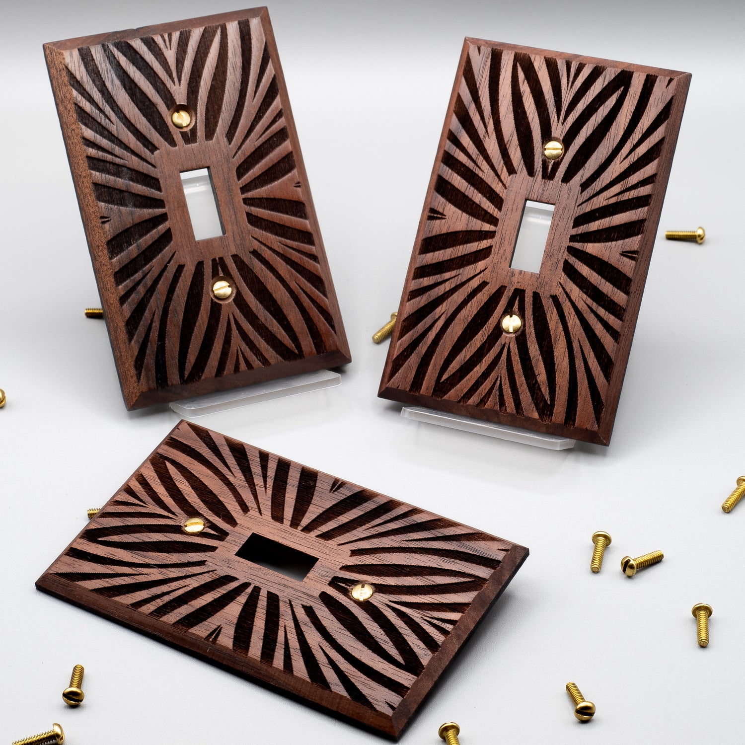 3 single toggle light switch plates made from Walnut wood on a white background surrounded by brass screws on all sides. The surfaces of the light switch plates are engraved with a Palm leaf pattern